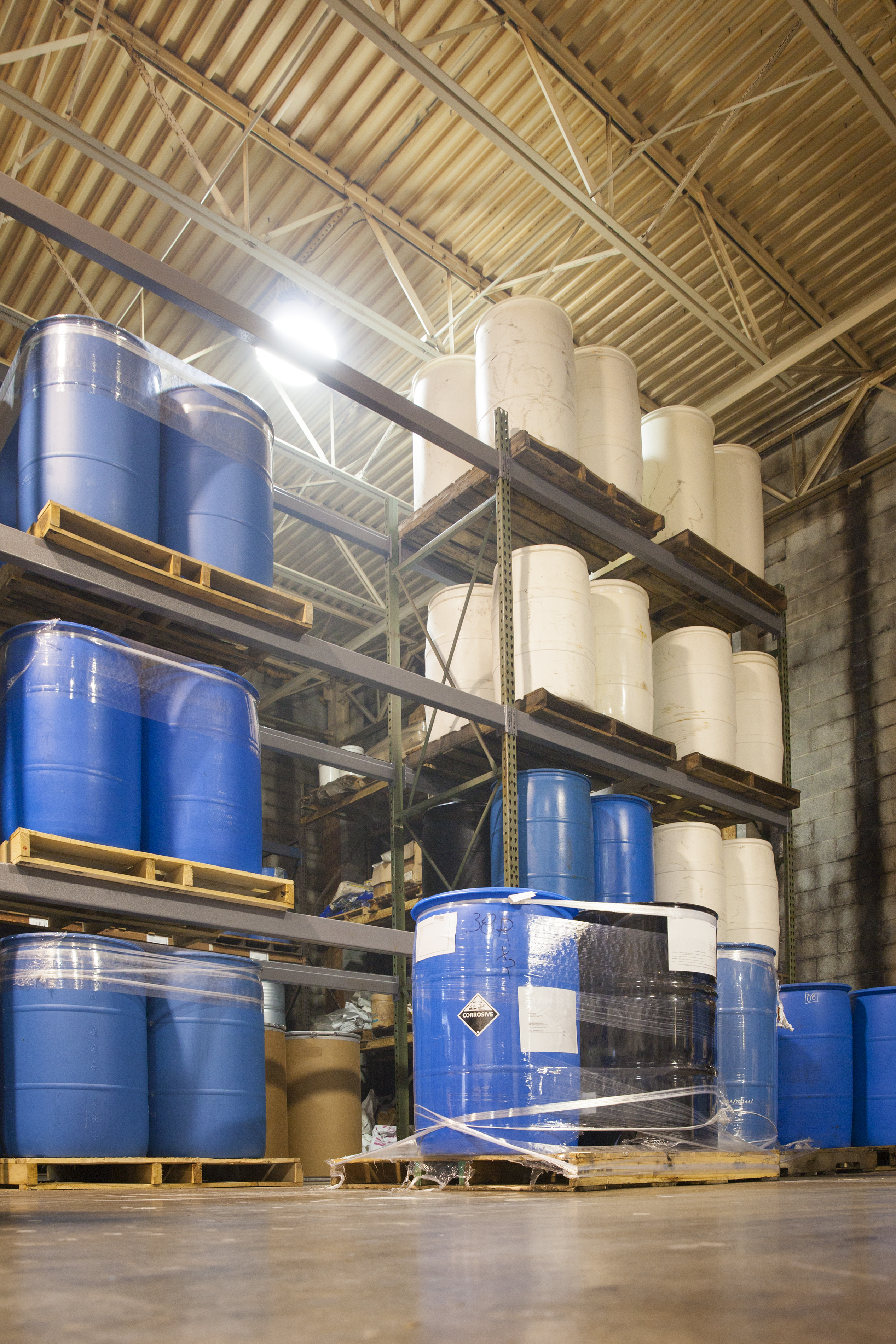 55 Gallon drums are stacked on pallets at an industrial chemical warehouse. A corrosive sticker is visible on one drum.
